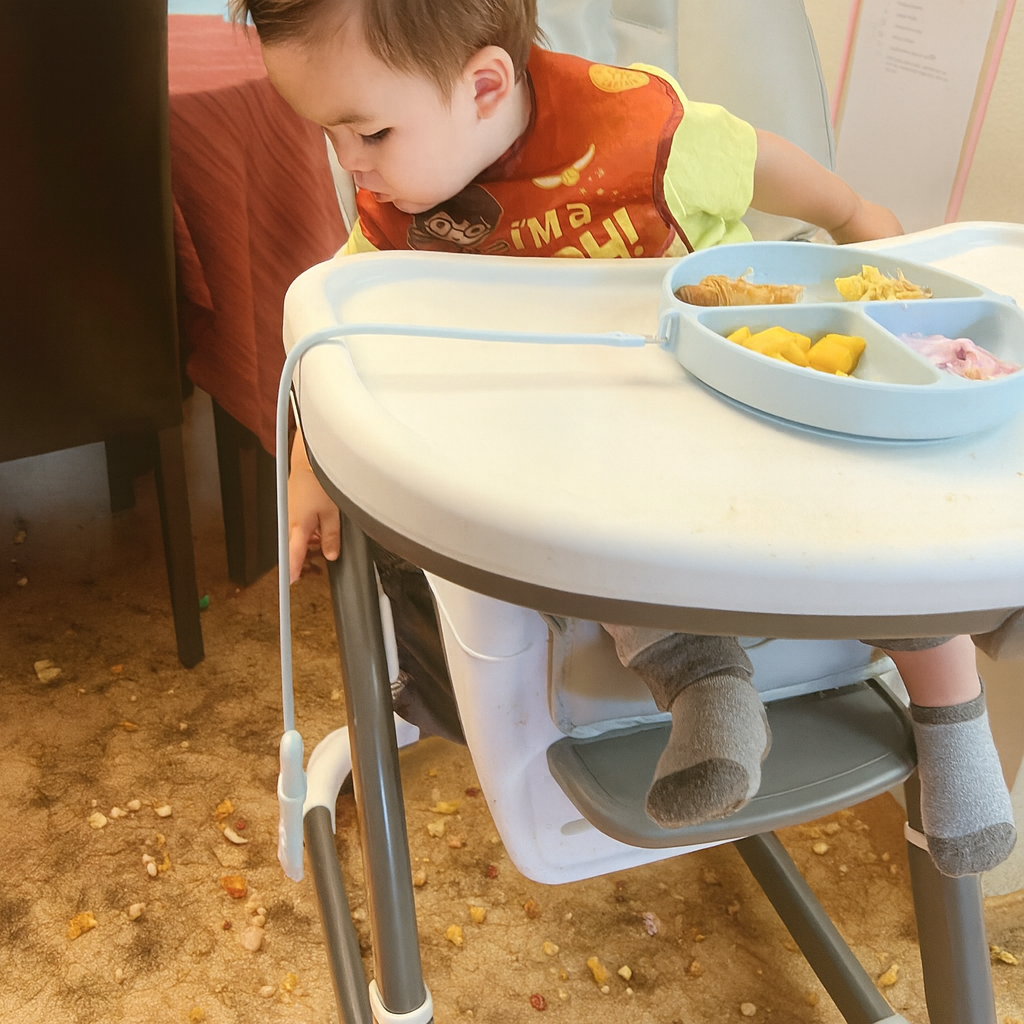 Child sitting in a high chair with a tray of food on a dirty floor.