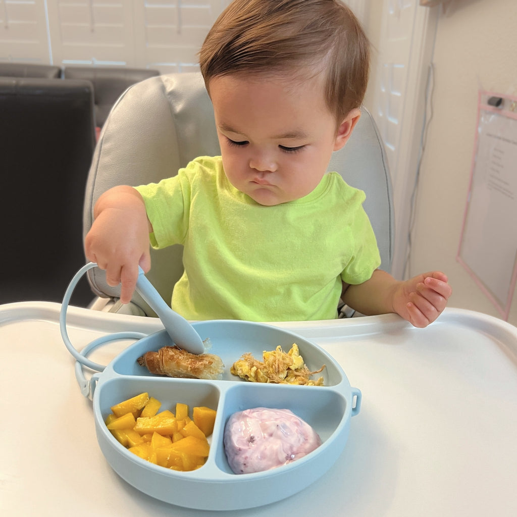 Child in a high chair eating from a divided baby plate with a spoon.