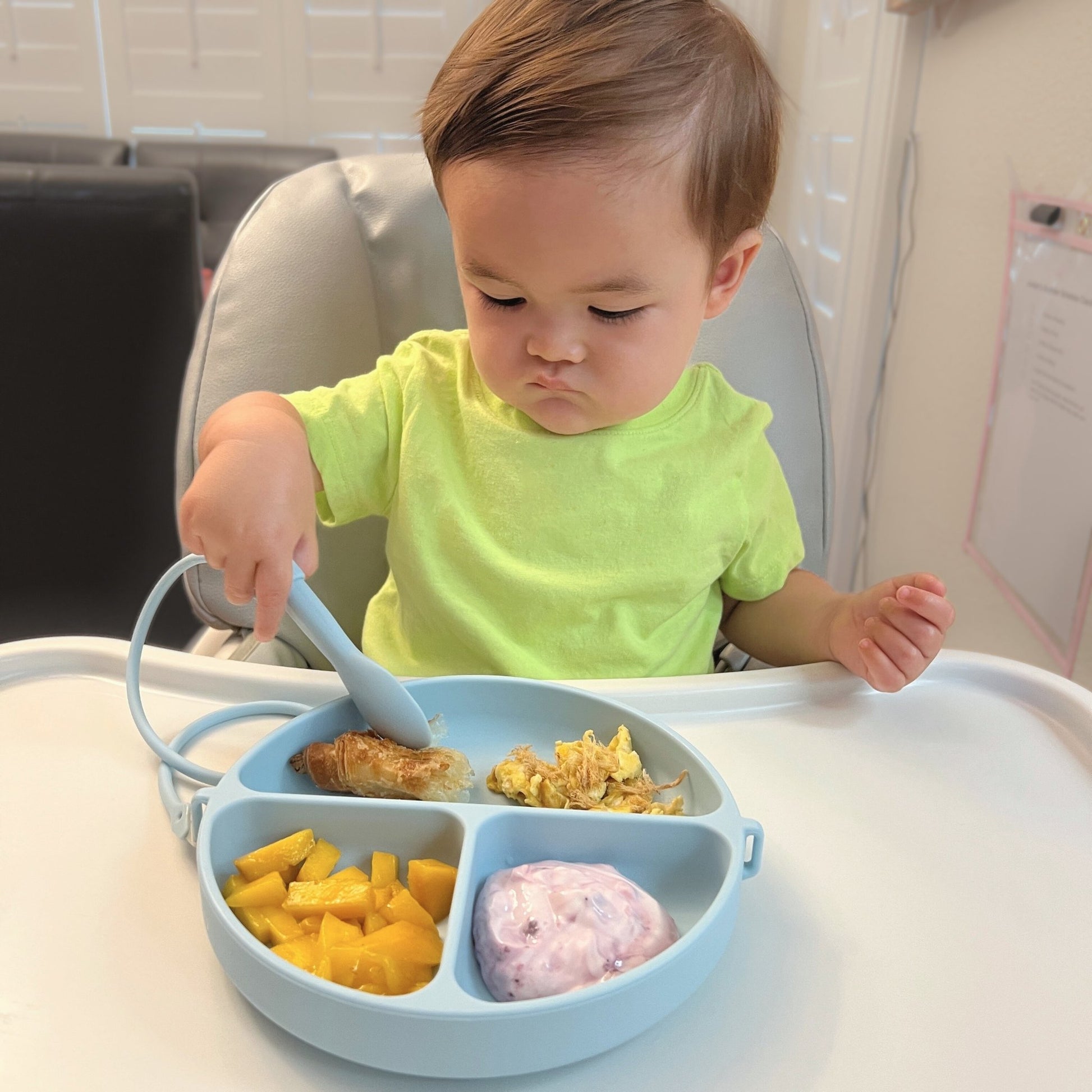Child in a high chair eating from a divided baby plate with a spoon.